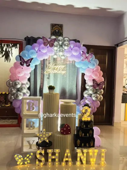 A butterfly-themed birthday setup perfect for a home celebration. A circular balloon arch in purple, pink, and silver creates a beautiful frame, complemented by light-up letters and 'BABY' blocks.