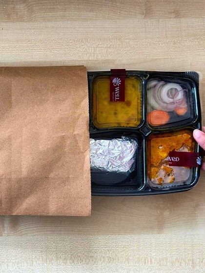Join me in unwrapping daily comfort, one home-style meal at a time. This image shows a packed thali being placed in a paper bag, ready for delivery.