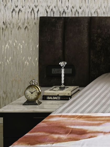 A close-up of the bedside details in a guest bedroom. A plush, dark brown headboard provides comfort, while the marble-topped nightstand offers a durable and elegant surface for essentials like an alarm clock and books.