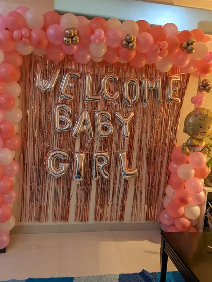 A beautiful 'Welcome Baby Girl' decoration for a home celebration, featuring a full balloon arch in shades of pink and white against a rose gold fringe curtain.