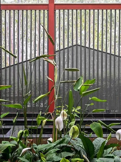 A close-up of the landscape design within the SARITHA SERENE office. The red structural beam cuts through the frame, contrasting with the delicate peace lilies and the vertical lines of the metal screen in the background.