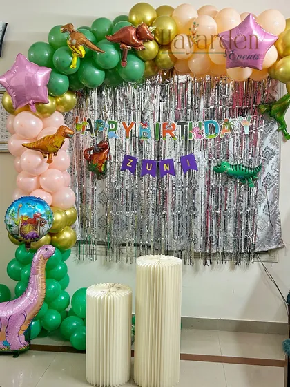 A dinosaur-themed birthday party with a green and peach balloon arch. Foil dinosaur balloons and a silver fringe curtain complete this prehistoric look.