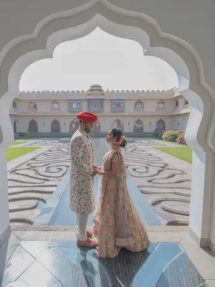 Framed perfectly within a large scalloped archway, a couple in regal wedding attire holds hands, looking out onto a magnificent palace courtyard. This shot uses symmetry and architecture to create a powerful, grand wedding portrait.