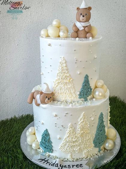 A cozy winterland-themed two-tier cake. This design features cute teddy bears, cream trees, and chocolate spheres to create a snowy, magical scene.