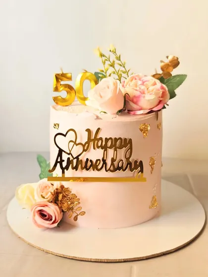 A beautiful golden anniversary cake with a simple design, adorned with pink flowers and a "Happy Anniversary" topper.