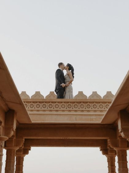 A dramatic and architectural portrait. The couple shares a kiss atop a stone structure at Suryagarh, creating a powerful silhouette against the sky that speaks of a love that is grand and timeless.