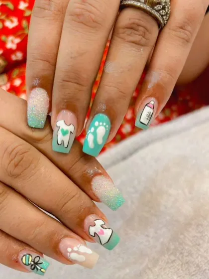 A cute baby shower-themed nail set with pastel green polish, glitter, and hand-painted baby items like bottles and footprints.