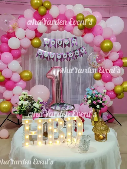 A simple and sweet first birthday setup with a balloon arch and 'HBD' marquee letters on the cake table.
