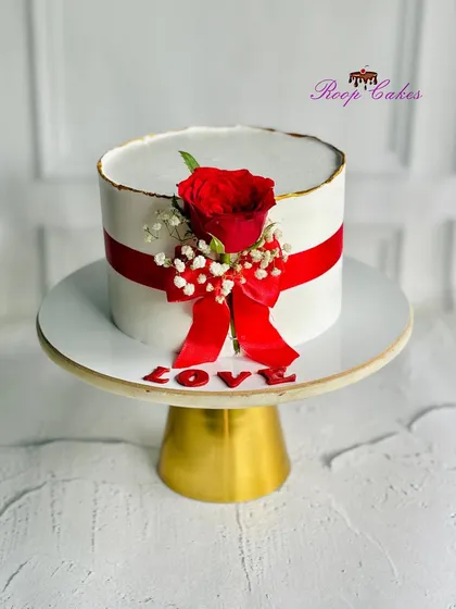 A sweet celebration of love. This elegant white cake is wrapped in a red ribbon and topped with a single red rose.