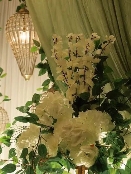 A detailed shot showing the interplay of light and texture. A crystal lantern illuminates the delicate white flowers and soft green drapery, creating a gentle and romantic ambiance.