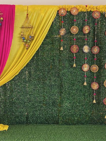 A modern Mehendi backdrop combining a lush green wall with bright pink and yellow drapes and traditional hangings.