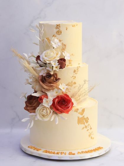 This three-tiered wedding cake captures the warmth of an autumn celebration. It's decorated with a cascade of handcrafted sugar flowers in rich, earthy tones of red, brown, and cream, complemented by flecks of edible gold.