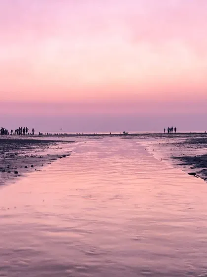 A channel of water on a Mumbai beach perfectly reflects the pink sky, leading the eye towards the people gathered at the shoreline.