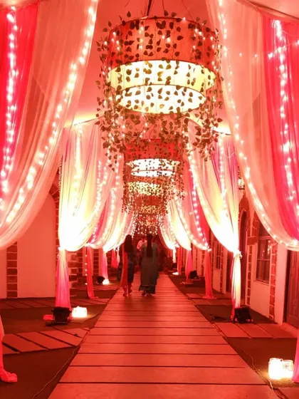 This entrance passage features large, custom-made chandeliers wrapped in vines, casting a warm, reddish glow. The walkway is lined with pink and white drapes and fairy lights, creating a rich and luxurious feel.