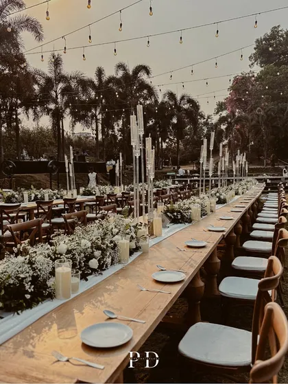 The long wooden tables adorned with floral runners and modern candelabras, set against the backdrop of palm trees and the setting sun.