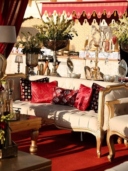 A luxurious lounge seating area where crimson and gold details pop against ivory furniture. The tablescape includes silver vases with red and white florals, elegant lamps, and patterned cushions to complete the royal look.