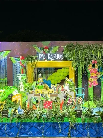 A vibrant neon jungle theme for a first birthday party. This wide shot shows the entire setup, filled with brightly colored animal props, lush greenery, and neon-style lighting effects.