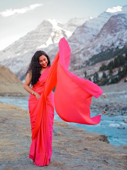 A solo shot of the bride, her pink saree flowing in the wind against the stunning mountain and river backdrop.