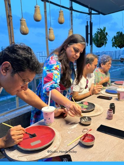 A couple and their family members are engrossed in painting their pottery pieces under the warm glow of the studio's hanging lights at dusk.