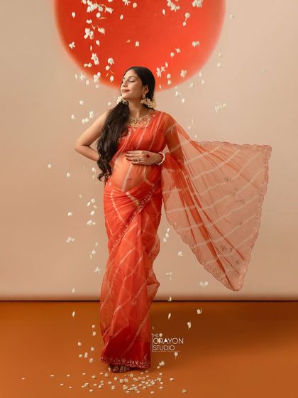 Draped in a vibrant orange organza saree, she stands in a shower of petals, embodying grace and warmth. This is a beautiful example of a fine art Indian maternity portrait.