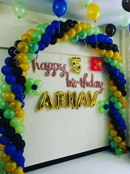 A simple and colorful balloon arch for a fifth birthday. Using a mix of green, blue, yellow, and black balloons, this archway decoration includes the name "ARNAV" and age in foil balloons.