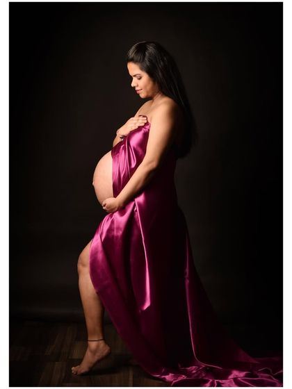 An artistic portrait using a single piece of magenta satin fabric draped elegantly around the body. The dramatic lighting highlights the curves of pregnancy beautifully.