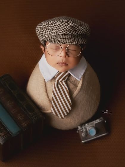 The little professor. This clever newborn portrait features a baby dressed in a tiny cap and glasses, posed with books and a miniature camera for a dapper, vintage look.