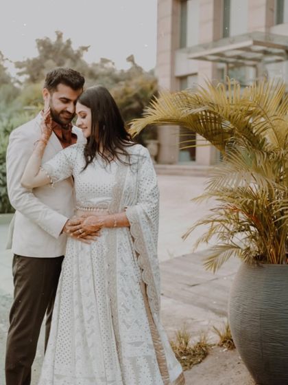 A romantic outdoor portrait of a couple in elegant white outfits, sharing a tender embrace in a modern, landscaped setting.