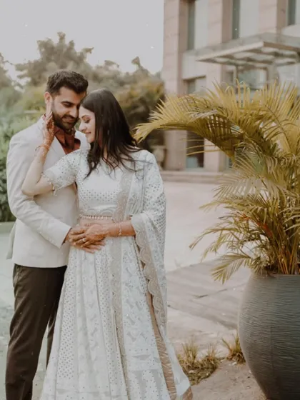 A romantic outdoor portrait of a couple in elegant white outfits, sharing a tender embrace in a modern, landscaped setting.
