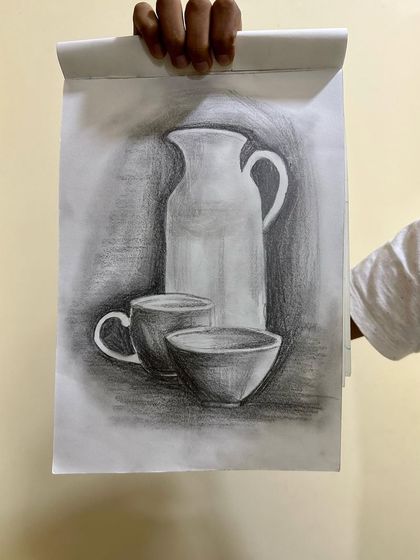 A student proudly holds up their still life drawing of a pitcher and cups.