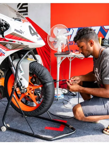 A mechanic works on a race bike in the pits, attaching tire warmers. This preparation is essential to ensure the tires are at optimal temperature for maximum grip right from the start of the race.