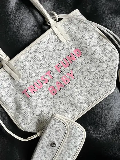 A cheeky "TRUST FUND BABY" design hand-painted in pink on a white Goyard purse and matching wallet.