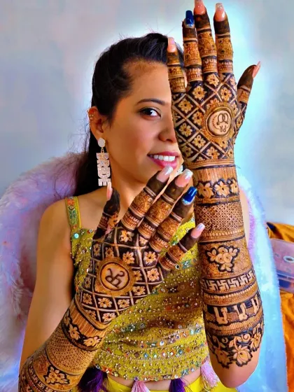 A happy bride showing off her full-hand engagement mehendi. The design features geometric patterns, floral bands, and the couple's initials, all coming together for a stunning bridal look.