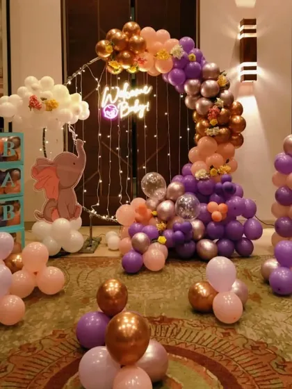 A beautiful baby shower setup with a 'Welcome Baby' neon sign. The decor features a balloon arch in pastel and chrome colors, cloud-like balloon clusters, and a cute elephant cutout, creating a warm and inviting atmosphere.