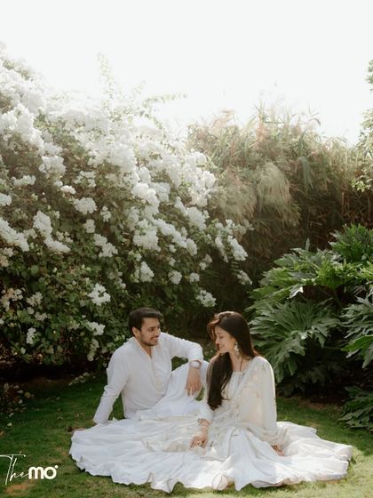 A serene pre-wedding moment. The couple, dressed in white, sits in a garden of white flowers, creating a peaceful and romantic image that feels pure and natural.