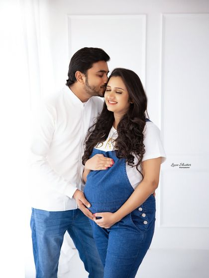 A sweet and casual maternity portrait. The couple's coordinated denim and white outfits create a fresh, modern look against a clean white studio background.