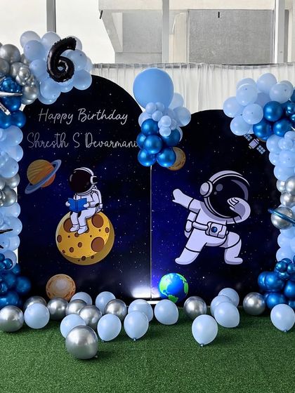 An awesome astronaut-themed setup for a 6th birthday. The backdrop shows astronauts in space, framed by a blue and silver balloon arch with planet and star accents.