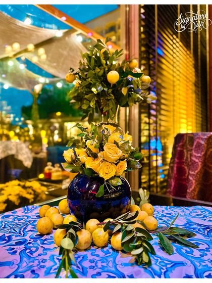 A beautiful tablescape featuring a miniature lemon tree and yellow roses in a blue vase. The intricate blue and white patterned tablecloth completes the Sicilian-inspired aesthetic.