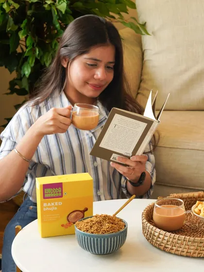A lifestyle image of a person enjoying baked bhujia with a cup of tea while reading, creating a cozy and relatable moment.
