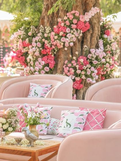 A beautiful 'Pichwai Haldi' setup in shades of pink. The tree is adorned with cascading pink flowers, and the seating features plush pink velvet sofas with custom-printed cushions, creating a royal and romantic ambiance.