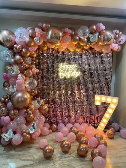 A 7th birthday celebration featuring a rose gold sequin wall, a pink and chrome balloon arch with butterfly details, and a light-up number '7'.