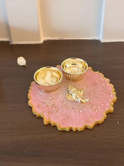 A pink resin thaali with a small Ganesha idol and gold leaf accents. This auspicious and aesthetically pleasing thaali is perfect for your Rakhi pooja.