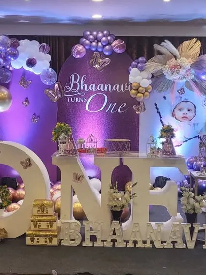 An elegant butterfly-themed first birthday celebration. The decor features a purple and gold color scheme, elaborate balloon arrangements, large "ONE" lettering for photos, and a beautifully decorated dessert table.