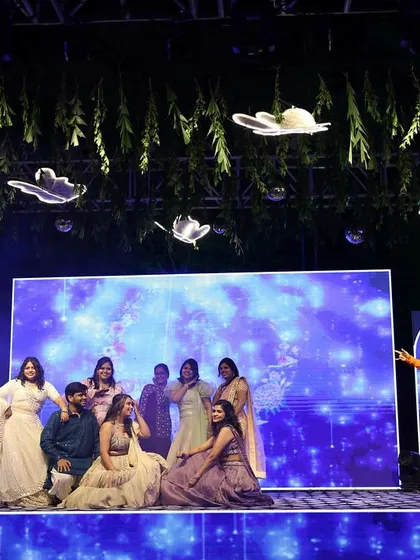 A wide view of the Sangeet stage with me hosting a family performance. The beautiful decor and lighting create a perfect backdrop for the celebrations.