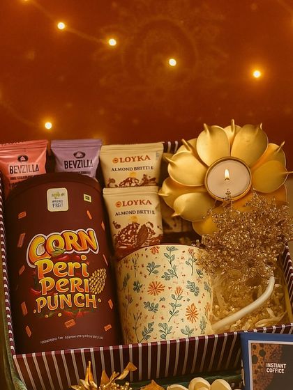 The Roshni Ka Tohfa, or 'gift of light', features a beautiful lotus-shaped brass diya as its centerpiece. It's complemented by a printed coffee mug, gourmet snacks, and coffee cubes, all arranged in a festive striped box.