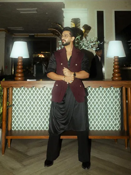 A client celebrating Diwali in a royal fusion look. He wears a black draped kurta set, layered with a double-breasted, sleeveless jacket in a rich burgundy, creating a look of modern royalty.
