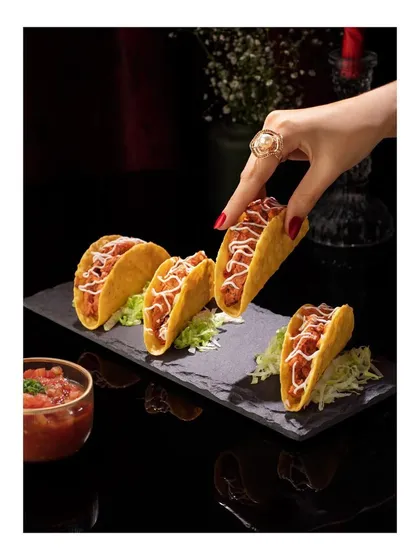 This image captures a hand picking up a crispy taco from a slate platter at Club Jolies. The dark, moody lighting and touch of human interaction create a chic and enticing scene for this popular dish.