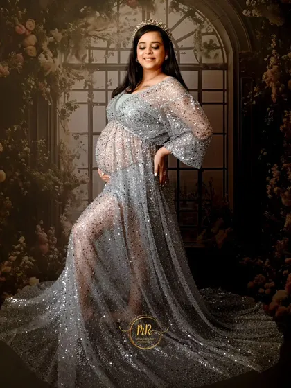 A glamorous solo maternity portrait. The mother-to-be glows in a sheer, sparkling silver gown, standing against a beautiful floral window backdrop in our studio.