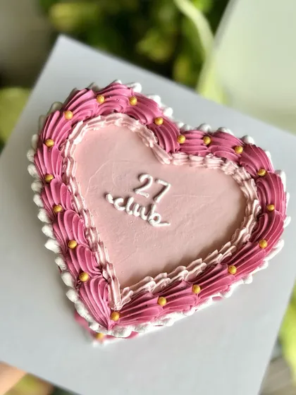 A top-down shot of the "27 club" heart cake, highlighting the perfect symmetry of the piped border and the delicate script in the center.