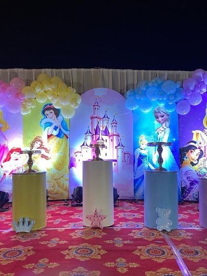 A grand princess-themed stage for a first birthday, featuring backdrops of various Disney princesses like Cinderella, Snow White, and Elsa, all tied together with colorful balloon arches.